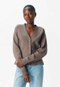 Brun cardigan med V-hals, fem knapper og ribbede manchetter. Båret over lyseblå jeans. Blød tekstur, afslappet pasform, minimalistisk design.