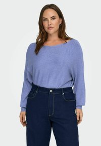 Helelelavendelvärviline, pikkade varrukatega topp soonilise tekstuuriga, kombineeritud tumeliste helesiniste kõrge vöökohaga teksadega, millel on kollane õmblus ja esitaskud.