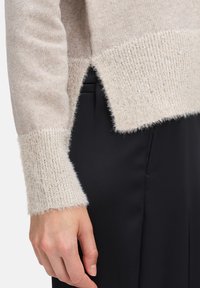 Jasnobeżowy sweter z dzianiny o puszystej fakturze i cekinowych ściągaczach. Posiada rozcięty dół oraz warstwowy fason, zestawiony z czarnymi spodniami.
