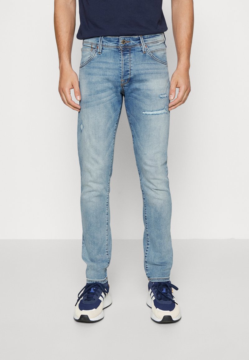 Άνδρας wearing ανοιχτό μπλε τζιν με φθορές και στενή γραμμή, με μπλε και λευκά αθλητικά παπούτσια σε ένα απλό λευκό φόντο.