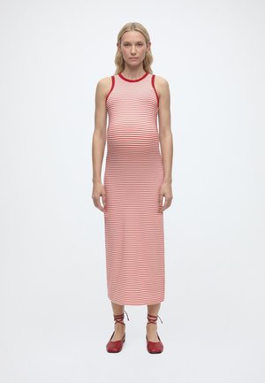 Femme blonde portant une robe midi sans manches à rayures rouges et blanches et des chaussures plates rouges à lacets, debout devant un fond blanc.