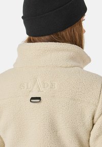 Giacca in pile color crema con colletto alto, caratterizzata dal logo ricamato "SLADE" e un dettaglio a loop nero sul retro.