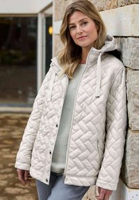 Helle, gesteppte Jacke in Beige mit Kapuze, die mit einem Rautenmuster, Druckknopfverschlüssen und einer Reißverschlusstasche ausgestattet ist, getragen über einem grauen Pullover.