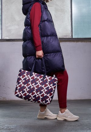 Personne en tenue bordeaux et veste matelassée bleu marine tenant un grand sac à motifs, portant des baskets beiges, debout à l'intérieur sur un sol en béton.