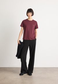 Woman with short bob haircut wearing a maroon t-shirt, black wide-leg pants, black sneakers, holding a black knit sweater, standing on a gray floor.