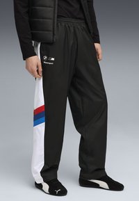 Sorte polyester træningsbukser med hvide sidepaneler, der har røde, blå og sorte striber, elastisk talje og BMW Motorsport-logo.