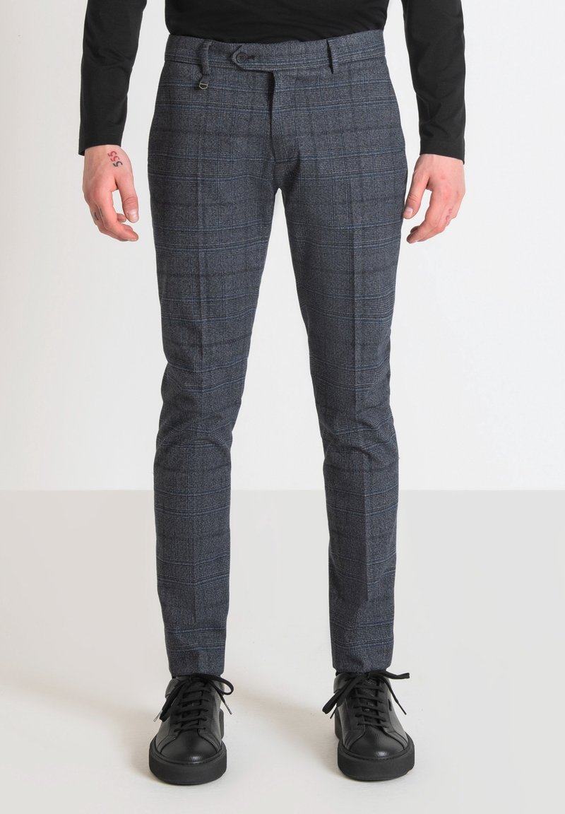 Homme portant un pantalon slim gris à carreaux et des baskets noires, debout devant un fond clair uni.