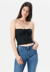 Top corto negro sin tirantes con detalle de nudo frontal, hecho de tela elástica; combinado con unos pantalones vaqueros azul de cintura alta.
