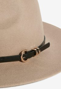 Béžový klobúk so širokou krempou z plsti s čiernou koženou páskou a zlatou okrúhlou prackou.