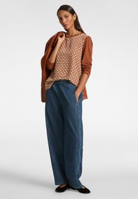 Maglietta marrone a maniche lunghe con fantasia sul petto, abbinata a jeans blu a gamba ampia con borchie laterali. Il modello posa con le mani nelle tasche.