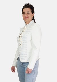 Chaqueta blanca ajustada con un cuello con volantes y textura de rayas verticales, que presenta botones dorados y un detalle de cremallera en contraste.