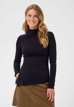 Jeune femme aux longs cheveux blonds ondulés porte un col roulé noir à manches longues et une jupe en velours côtelé marron, souriante, la main dans la poche.