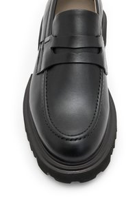 Mocassin en cuir noir avec un bout mocassin, présentant une sangle décorative sur le dessus et une semelle en caoutchouc texturé.