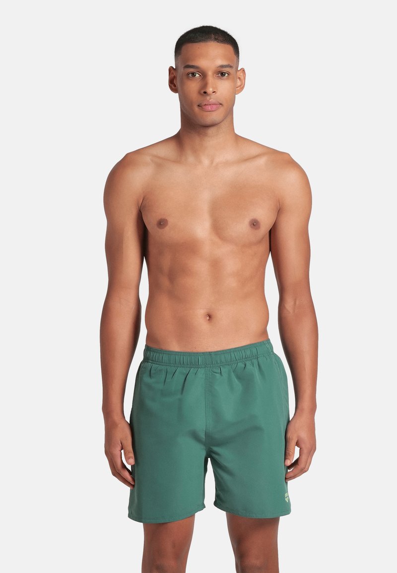 Uomo a torso nudo che sta in piedi frontale indossando un costume da bagno verde con vita elasticizzata su uno sfondo bianco uniforme.