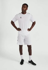 Camiseta deportiva blanca y pantalones cortos con acentos y logotipo negros, combinados con calzado negro y calcetines blancos, de pie frente a un fondo liso.