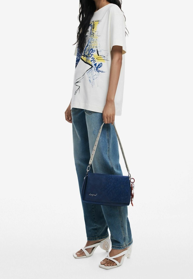 Femme portant un t-shirt blanc avec un motif graphique et un jean bleu, tenant un sac à main bleu avec une bandoulière à motif, portant des sandales blanches à talons.