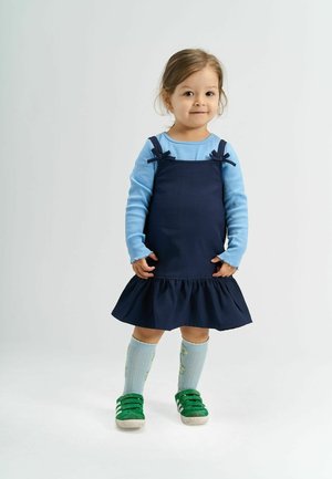 Rochie tip salopetă de culoare albastru marin, cu tiv ondulat, asortată cu un tricou cu mânecă lungă de culoare bleu deschis și pantofi verzi, accentuată de șosete cu model floral.