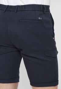 Bermudas en coton bleu marine avec un design à l'avant plat, une poche arrière et un ourlet roulé. Présente un subtil détail de logo près de la taille et de la poche.