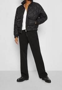 Czarna pikowana kurtka bomberka z dwoma przednimi kieszeniami, ściągaczami przy rękawach i wysokim kołnierzem, noszona na białej bluzce i zestawiona z luźnymi czarnymi spodniami.