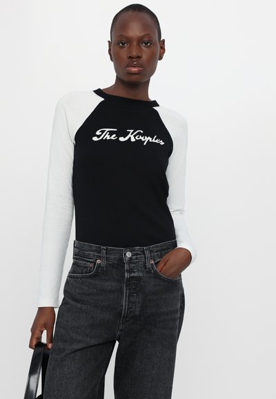 Camisa de manga larga en blanco y negro con un diseño raglán que presenta "The Karpules" en una escritura blanca. Combinada con unos jeans de mezclilla oscuros de tiro alto.