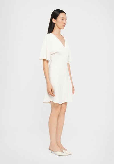 Witte jurk met een V-hals, korte fluttermouwen en een uitlopende zoom. Gladde, gestructureerde stof. Gecombineerd met crème slingback schoenen.