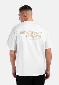 Hvid kortærmet t-shirt med en glat tekstur, rund halsudskæring og teksten "elevate your Everyday" i beige på ryggen.