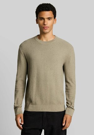 Giovane uomo in piedi che indossa un maglione beige a collo tondo con trama e pantaloni neri su uno sfondo grigio uniforme.