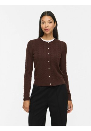 Femme aux longs cheveux foncés portant un cardigan marron en maille torsadée boutonné et un pantalon noir, debout devant un fond blanc uni.
