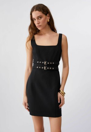 Giovane donna con abito nero senza maniche e scollo quadrato, cintura doppia con fibbie, orecchini dorati e braccialetti su sfondo neutro.