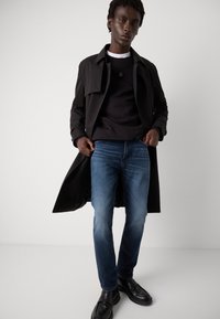 Abrigo largo negro, sudadera negra, jeans azules y zapatos negros. El conjunto presenta un diseño limpio con un ajuste relajado y texturas contrastantes.