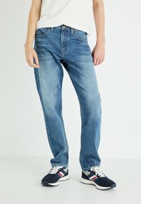 Jeans de denim azul claro com um corte descontraído, apresentando um fecho de botão e bolsos frontais, usados com ténis azul-marinho e uma camisa branca.
