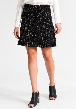Mujer con falda negra con lunares blancos, blusa blanca de manga larga y zapatos negros de tacón abiertos, de pie sobre fondo blanco.