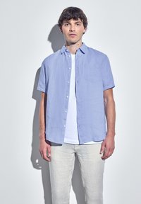 Camicetta azzurra a maniche corte con bottoni e taschino sul petto, indossata sopra una maglietta bianca, abbinata a pantaloni grigi chiari.