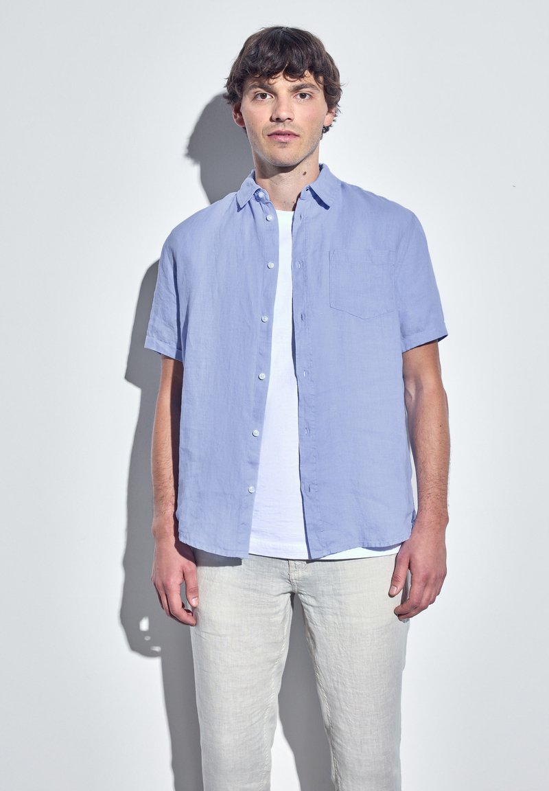 Camicetta azzurra a maniche corte con bottoni e taschino sul petto, indossata sopra una maglietta bianca, abbinata a pantaloni grigi chiari.