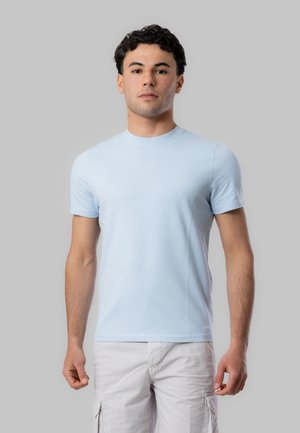 Giovane uomo con capelli corti, ricci e scuri, che indossa una t-shirt azzurra a maniche corte e pantaloni cargo bianchi, in piedi davanti a uno sfondo grigio.