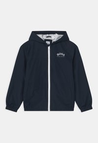Chaqueta cortavientos azul marino con capucha, con cierre frontal de cremallera, logo blanco en el pecho y puños y dobladillo elásticos.