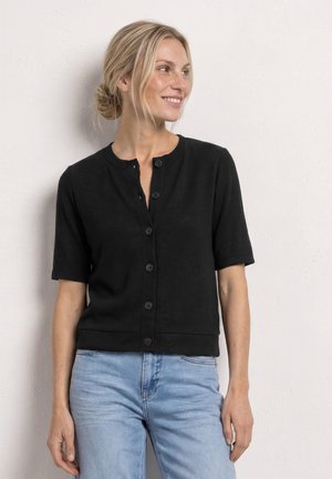 Lachende vrouw met blond haar in een lage knot, die een zwart knoopjeshemd met korte mouwen en lichtblauwe spijkerbroek draagt, staande tegen een witte muur.