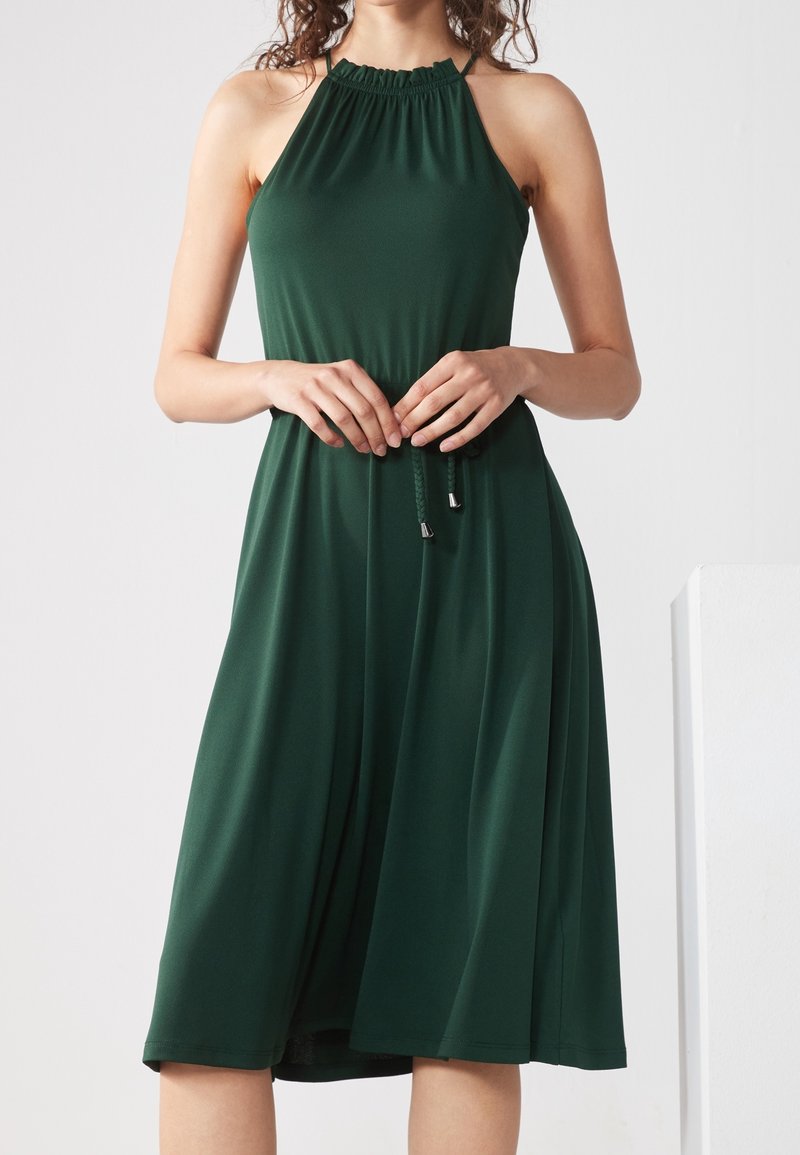 Femme portant une robe midi sans manches vert foncé avec un col montant froncé et une ceinture tressée à la taille, debout devant un fond uni.