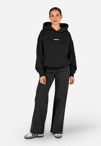 Crna dukserica s kapuljačom i prednjim džepom, s bijelim logotipom, upairana s crnim širokim trapericama. Materijali izgledaju mekani, s ležernim dizajnom.