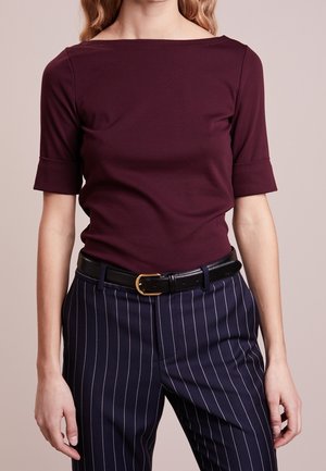 Vrouw met een bordeauxrode top met korte mouwen en marineblauwe gestreepte broek met een zwarte riem en gouden gesp tegen een effen achtergrond.