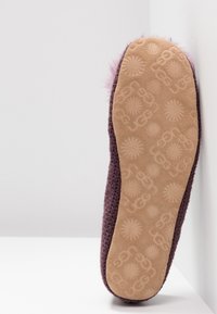 Podeszwa fioletowego wełnianego kapcia z brązową gumową podeszwą, zdobioną wzorami promieni słonecznych i łańcuchami, oparta o białe narożne ściany.