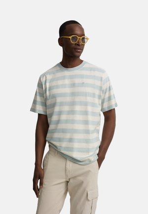 Hombre con camiseta de rayas azul pálido y blanca, pantalones cargo beige y gafas redondas amarillas, de pie con una mano en el bolsillo.