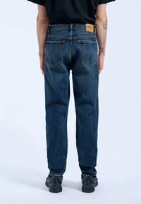 Jeans de denim azul oscuro con corte recto, cinco bolsillos y una etiqueta de cuero en la parte posterior de la cinturilla. Costuras visibles y un acabado texturizado.