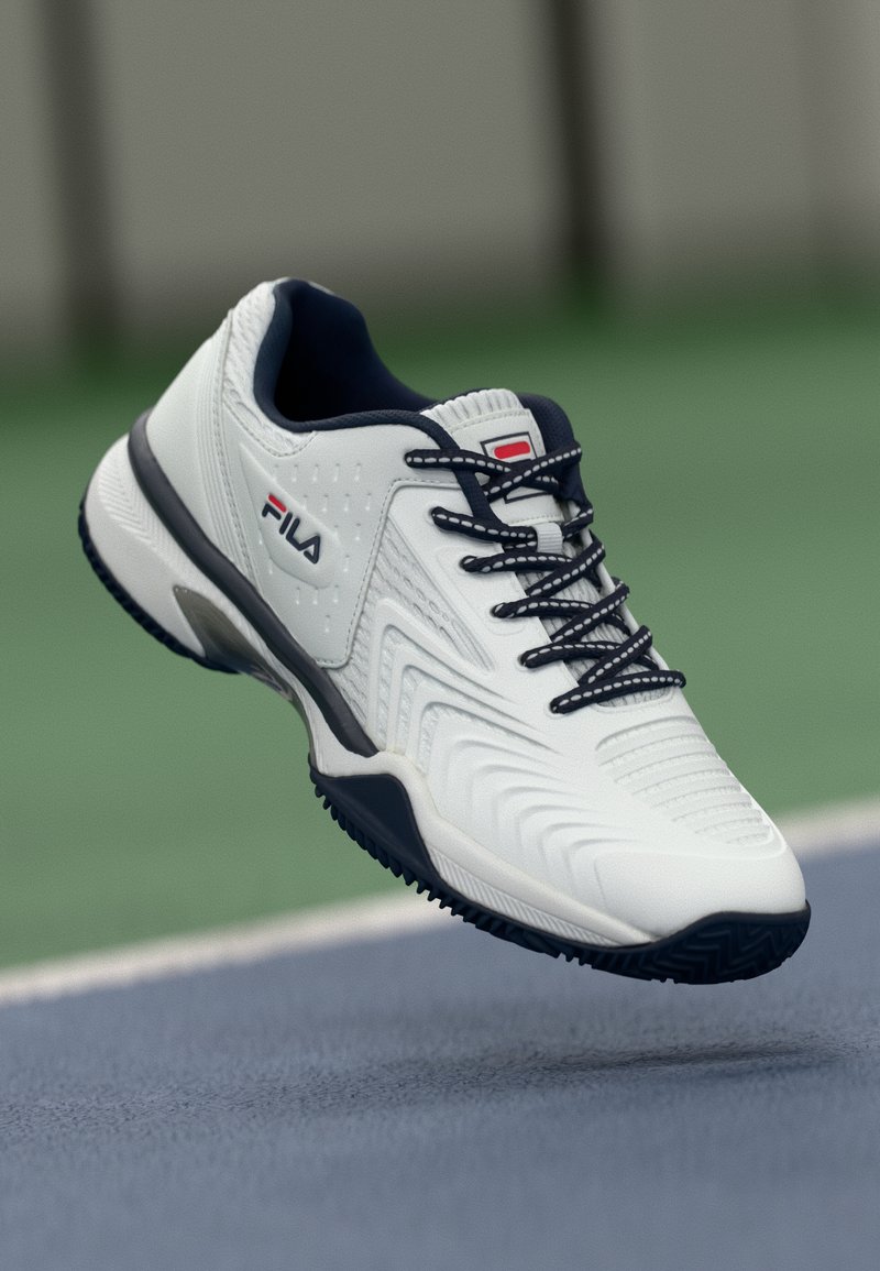 Biele športové tenisky s textúrovaným zvrškom, námorníckymi akcentmi a čiernou gumovou podrážkou. Obsahujú dizajn na šnurovanie a logo Fila na boku.