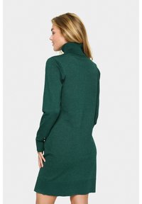 Mujer con cabello rubio vestida con un vestido de suéter de cuello alto de manga larga en verde oscuro, de pie de lado contra un fondo blanco liso.