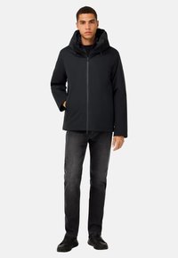 Melnas kapuces jaka ar rāvējslēdzēju priekšā, sānu kabatām un gludu, maigu tekstūru; savienota ar tumšiem džinsiem un melniem apaviem.