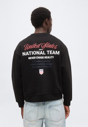 Homme de dos portant un sweat-shirt noir avec "United States Men's National Team" et un texte motivant en rouge, blanc et bleu dans le dos.