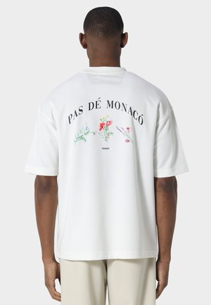 Uomo che indossa una T-shirt bianca oversize con la scritta "PAS DÉ MONACÔ" e piccole illustrazioni colorate di fiori sul retro, abbinata a pantaloni chiari.