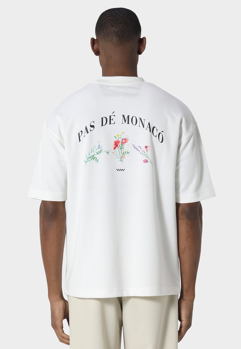 Uomo che indossa una T-shirt bianca oversize con la scritta "PAS DÉ MONACÔ" e piccole illustrazioni colorate di fiori sul retro, abbinata a pantaloni chiari.