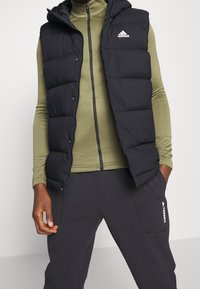 Osoba v černé péřové vestě Adidas, olivově zeleném dlouhém rukávu s zipem a černých kalhotách Adidas Terrex s rukama v kapsách.